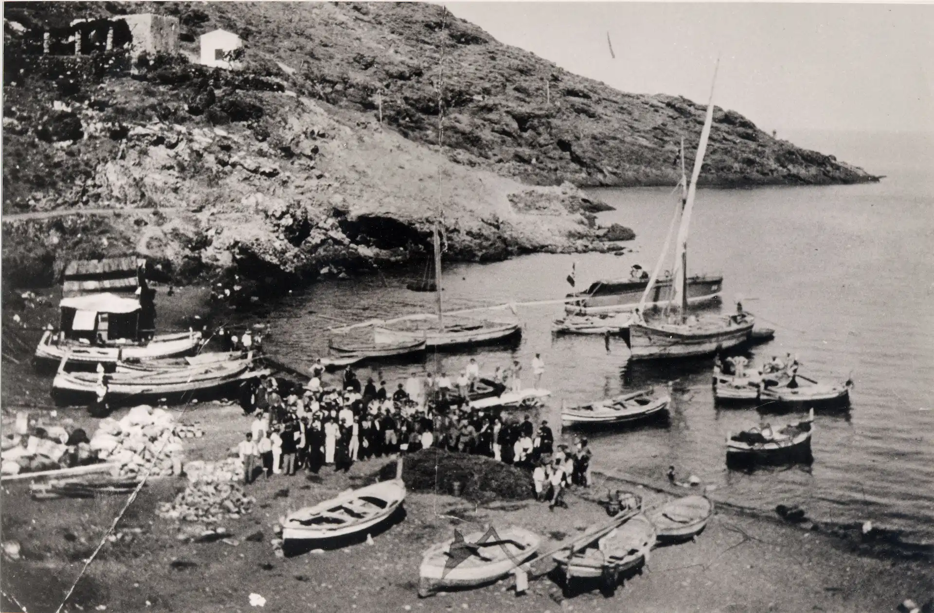 Passeggeri affollati sulla spiaggia in attesa di imbarcarsi. Un pontile di legno mobile (un piano inclinato di legno su due ruote con un lato sull’asciutto della sabbia) consentiva con un saltello di salire sulla barca a remi che li porterà sotto bordo alla nave all'ancora al centro della cala; dalla barca accostata sul fianco della nave si saliva a bordo utilizzando una scaletta mobile.Un’operazione complessa che impegnava i marinai in servizio sulla barca